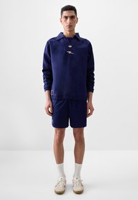 Mörkblå sweatshirt med hög krage, vit adidas-logotyp, i kombination med mörkblå randiga shorts och krämfärgade sneakers med röda accenter. Vita strumpor.