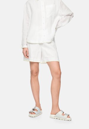 Frau trägt ein weißes, langärmliges Hemd mit Knöpfen und Tasche, weiße Shorts und weiße Slip-on-Sandalen mit großen dekorativen Schnallen.