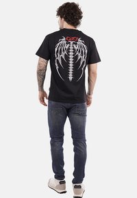 T-shirt nera a maniche corte con una grafica bianca di spina e ali sul retro, indossata con jeans blu e sneakers chiare.