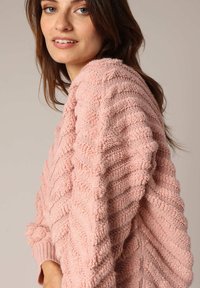 Pull en maille texturée rose clair avec un motif en zigzag et des poignets côtelés. Le tissu semble doux et a une sensation pelucheuse.