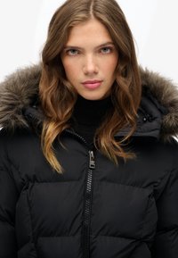 Giacca a vento nera con cappuccio bordato in pelliccia, dotata di chiusura con zip e design trapuntato. Il tessuto sembra liscio e isolato.