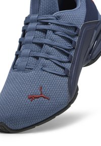 Zapatilla deportiva de malla azul con parte superior texturizada, talón reforzado, suela negra y logo bordado rojo. Presenta un sistema de cordones elásticos para un ajuste seguro.