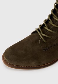 Botte en daim vert olive foncé à lacets avec semelle en caoutchouc marron et coutures détaillées sur le côté et la zone des orteils.
