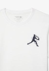 Camiseta de algodón blanca con una silueta en azul marino de un jugador en movimiento en la parte frontal. Diseño de corte regular con cuello redondo.