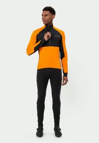 Tricou sport pentru bărbați cu mâneci lungi, de culoare portocalie și neagră, fabricat din material elastic, având un guler cu fermoar și o siluetă ajustată. Pantalonii negri completează ținuta.