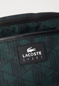 Žalia raštuota rankinė, pagaminta iš tekstūruotos medžiagos, su juodu užtrauktuku ir stačiakampiu Lacoste Sport logotipo emblemą.