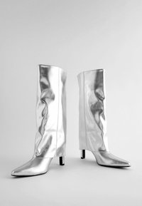 Bottes montantes en métal argenté avec un bout pointu, un design épuré et des talons carrés. Texture lisse avec des détails en tissu froncé.