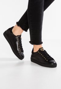 Svarta lädersneakers med guldöglor, texturerade svarta hälaccenter, rund tå och platt svart sula, kombinerade med trasiga svarta jeans.