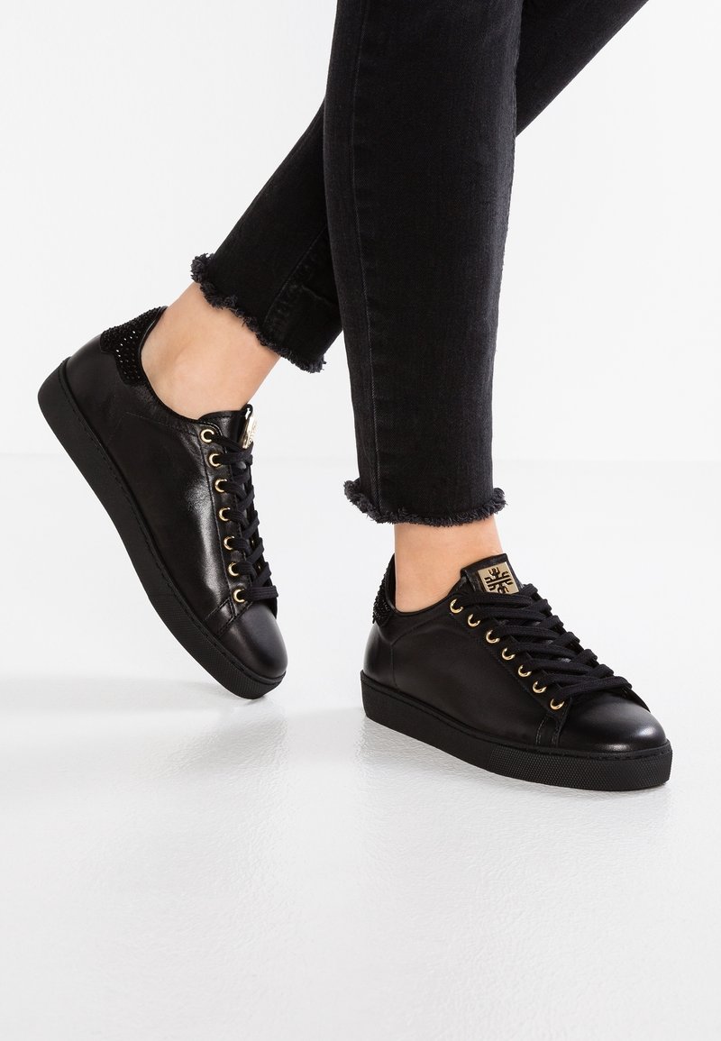 Svarta lädersneakers med guldöglor, texturerade svarta hälaccenter, rund tå och platt svart sula, kombinerade med trasiga svarta jeans.