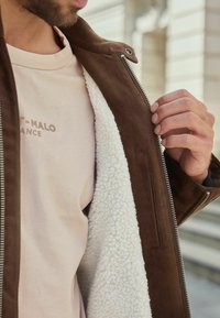 Uomo che indossa una camicia beige mentre apre la giacca marrone con rivestimento in pelliccia bianca, mostrando i dettagli della cerniera e delle trame del tessuto.