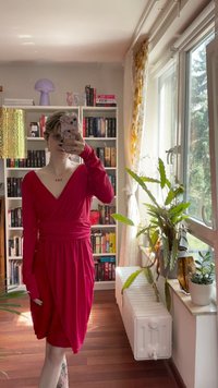 Czerwona sukienka o kroju kopertowym z długimi rękawami i głębokim dekoltem w kształcie V. Tkanina miękko opada. W tle widoczne są regały z książkami i rośliny doniczkowe.
