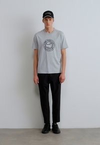 Pilkos spalvos marškinėliai su teniso grafika, juodos, laisvo stiliaus kelnės ir juodi sportbačiai; dėvi juodą kepurę su balta logotipu. Vienos spalvos fonas.