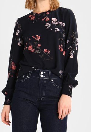 Blouse à manches longues florale en bleu marine avec des motifs roses et blancs, col rond et poignets élastiques. Associée à un jean en denim foncé taille haute.