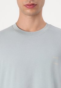 T-shirt en coton bleu clair avec un col rond et un subtil imprimé doré du logo de la marque sur le côté gauche. Texture lisse avec une coupe classique.