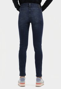 Dunkelblaue Skinny-Jeans mit glatter Textur, zwei Gesäßtaschen und einem Lederpatch am Taillenbund, kombiniert mit hellgrauen Sneakern.