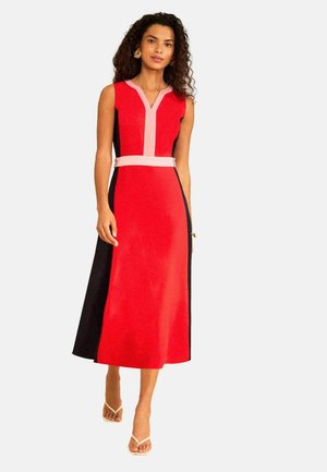 Göndör hajú nő piros, ujjatlan midi ruhában, rózsaszín szegéllyel és fekete oldalpanelekkel, előrefelé sétálva fehér szandálban.