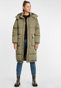 Olivgrön puffjacka med avtagbar huva, framknäppning med tryckknappar och dragkedja, stora fickor och elastiska muddar. Bärs med blå jeans och stövlar.