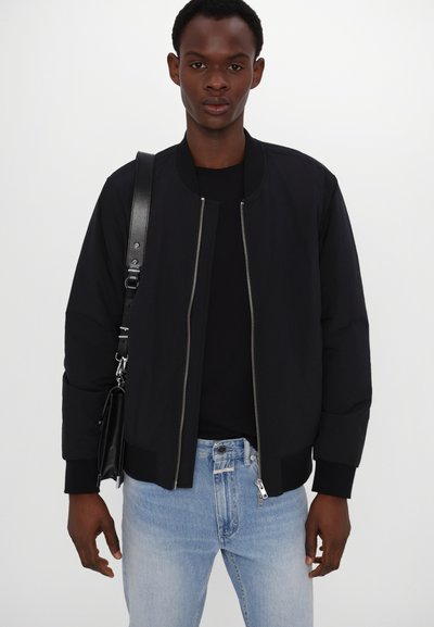 Veste bomber noire avec des poignets et un col côtelés, fermeture éclair à l'avant, assortie à un t-shirt noir et un jean bleu clair, tenue avec une sangle de sac noire.