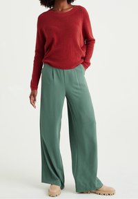 Ruskean värinen neulepaita, jossa on pyöreä pääntie, yhdistettynä väljiin vihreisiin leveälahkeisiin housuihin ja beigekirjaisiin korkokenkiin. Yhteensopiva pinta on sileä.