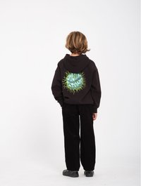 Sudadera negra con diseño gráfico verde en la parte de atrás, combinada con pantalones de pana negros y zapatos negros. Fondo blanco liso.