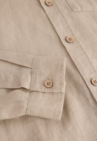 Detail eines beigen Leinenhemdes mit einem umgeschlagenen Ärmelbündchen mit einem einzelnen Knopf und einer geknöpften Knopfleiste, das die natürliche Struktur und Verarbeitung des Stoffs hervorhebt.