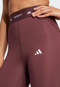 Leggings de sport bordeaux avec une large taille comportant le texte "TECHFIT" et des accents de logo Adidas blancs. Tissu lisse et extensible.