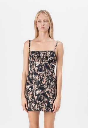 Femeie cu păr blond lung, purtând o rochie mini mulată, fără mâneci, cu un model floral abstract negru și bej, stând în fața unui fundal alb.