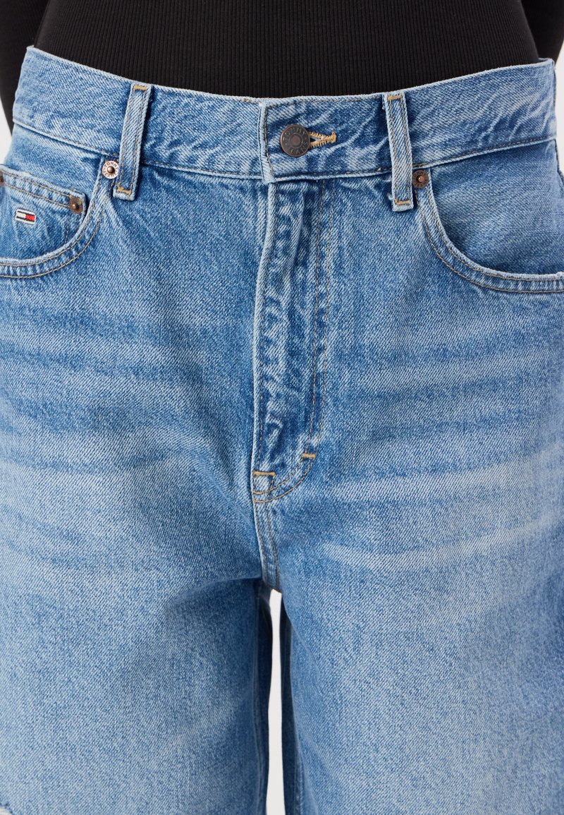 Shorts en jean bleu avec taille haute, comportant une fermeture par bouton et une décoloration subtile. Inclut des poches avant et une petite étiquette de logo.