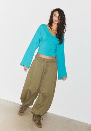 Femme aux longs cheveux bouclés portant un haut turquoise à manches longues, un pantalon ample vert olive et des baskets montantes marron.
