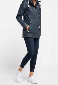 Imperméable bleu marine avec capuche, accents blancs aux fermetures à bascule, boutons-pression et poches latérales. Associé à un jean foncé et des baskets blanches.