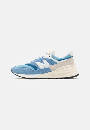 Sneaker azzurro chiaro con texture diverse in suede e mesh, accento di logo bianco, suola imbottita e un dettaglio sul tallone grigio in contrasto.