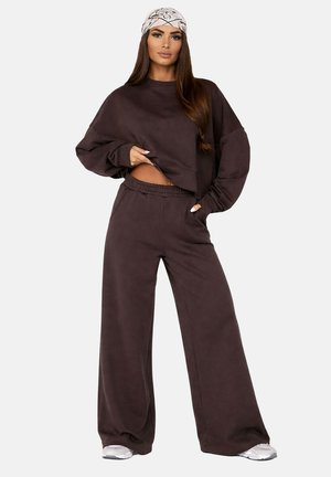 Femme portant un sweat-shirt ample marron foncé et un pantalon à jambes larges, des baskets blanches et un foulard blanc à motifs, debout devant un fond uni.