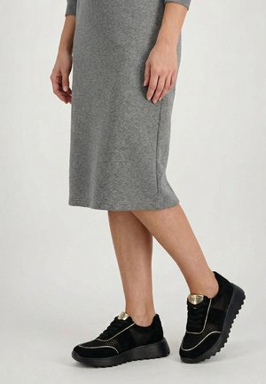 Kobieta ubrana w szarą sukienkę do kolan i czarne sportowe buty z siateczki ze złotymi akcentami, stojąca na tle jasnego, jednolitego tła.