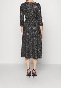 Robe midi noire et pailletée avec des manches trois-quarts, une taille cintrée et une jupe évasée. Portée avec des sandales à talons hauts noires à lanières.