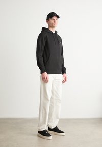 Pulover cu glugă negru, cu buzunar frontal și șnur, asociat cu pantaloni largi albi și adidași din suede negri cu talpă crem.