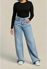 Personne portant un jean taille haute bleu clair à jambes larges, un haut noir à manches longues, des baskets blanches, debout avec une main dans la poche.