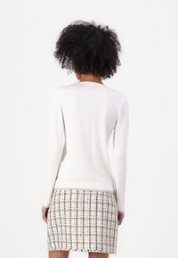 Haut blanc à manches longues avec un col rond et une texture lisse, associé à une jupe en tweed multicolore avec un motif à carreaux et des bords effilochés.