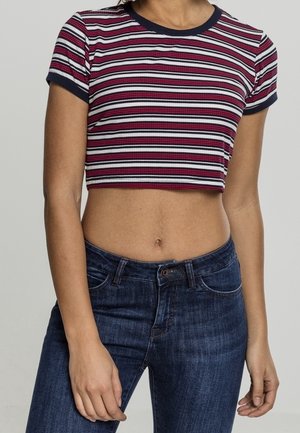 Osoba ubrana w czerwoną, białą i granatową pasiaste, krótki rękaw, krótki top oraz ciemnoniebieskie, wysokowalne dżinsy, stojąca z ramionami rozluźnionymi.