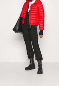 Chaqueta acolchada roja con capucha, cierre de cremallera y cordones ajustables; pantalones cargo negros; botas negras con cordones; bolso tote grande negro.