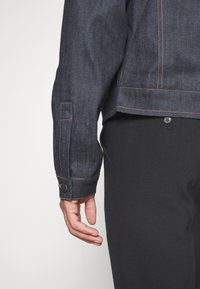 Primer plano de una mano que lleva una chaqueta de mezclilla azul oscuro con costuras beige y pantalones negros con un botón visible en el bolsillo trasero.