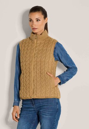 Frau trägt eine beige, kabelgestrickte Weste mit Reißverschluss über einem blauen Jeanshemd und blauen Jeans, sie steht mit einer Hand in der Westentasche.