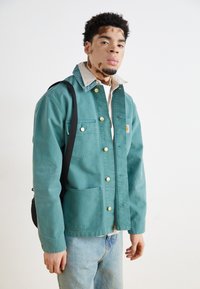 Veste en toile turquoise avec des boutons-pression dorés, poches avant et col beige, assortie à un jean bleu clair et un sac à bandoulière noir.
