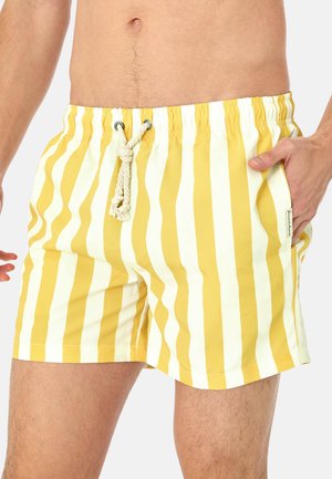 Mand iført gule og hvide lodrette stribede badeshorts med snøre i taljen, hånd i lommen, foran en ensfarvet baggrund.