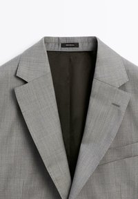 Hellgrauer Blazer mit fallendem Revers, einer Brusttasche und glattem Innenfutter. Der Stoff zeigt eine dezente Webstruktur. Das Etikett trägt die Aufschrift "Made in Portugal."