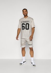 Γκρι αθλητική ενδυμασία που περιλαμβάνει ένα πλέγμα μπλουζάκι με κατακόρυφες ρίγες, μαύρα γράμματα "CHIEFS" και σορτσάκια με αντίστοιχη σχεδίαση και άσπρες λεπτομέρειες.