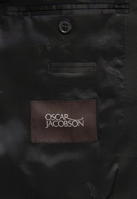 Zwart satijnen stof heeft een knoop en een zak met een bruine label dat in witte draad "OSCAR JACOBSON" is geperst.