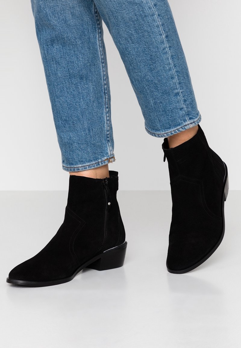 Osoba w czarnych zamszowych botkach na kostkę z bocznym zamkiem i niebieskich krótkich dżinsach na jasnoszarym podłożu.