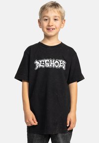 Černé oversized tričko vyrobené z bavlny, s bílým grafickým textem "DESHOES" na přední straně. Krátké rukávy a kulatý výstřih.