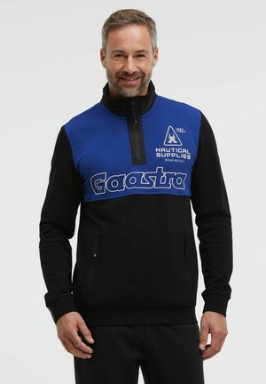 Mann mit kurzem Haar und Bart, der einen blau-schwarzen Gaastra-Pullover mit dem Text "Nautical Supplies" und Fronttaschen trägt, lächelnd vor einem einfarbigen Hintergrund.