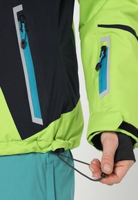 Světlá zelená a černá bunda s kapsami na zip, šedými detaily a nastavovatelným spodním lemem. Má elastické manžety a hladkou texturu.